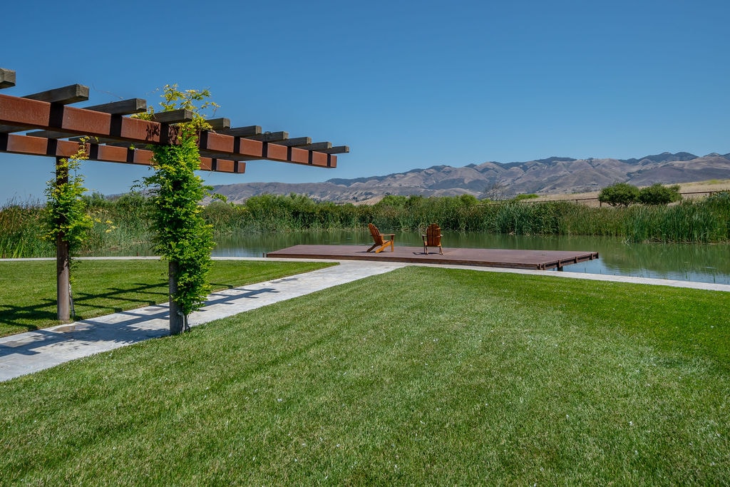 Chairs on a pond-side deck beside a lawn and pergola